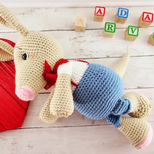 Può includere: Un peluche di peluche a forma di canguro, sdraiato su una superficie di legno bianco. Il canguro è beige con il naso rosa e un fiocco rosso. Indossa una camicia a righe blu e bianche. Il canguro è circondato da blocchi di legno con lettere, che compongono la parola "ADARVRK".