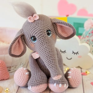May include: A crocheted gray elephant toy with pink ears, a pink bow, and a gray tail. The elephant has blue eyes and a pink nose. The toy is sitting on a white wooden surface.