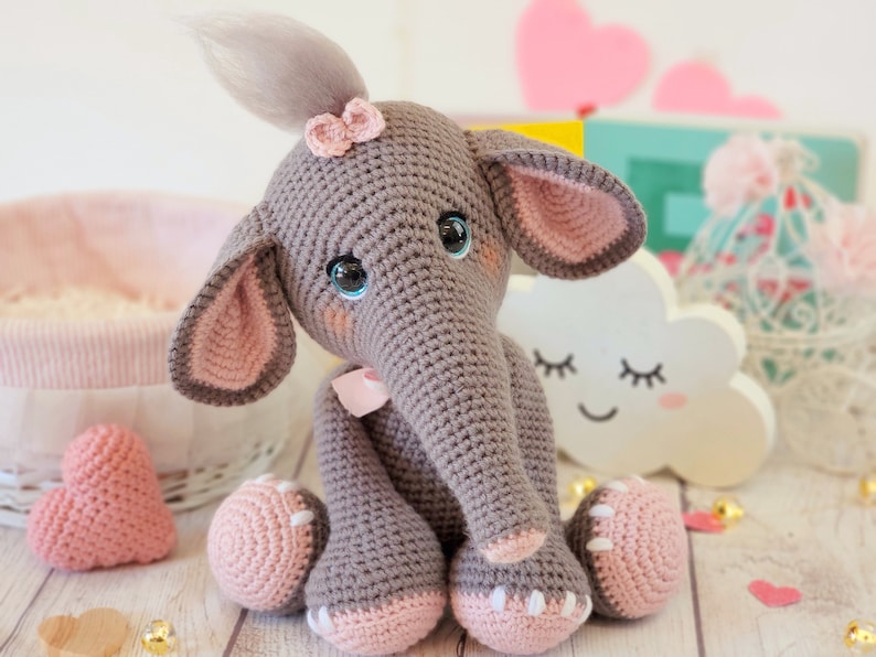 May include: A crocheted gray elephant toy with pink ears, a pink bow, and a gray tail. The elephant has blue eyes and a pink nose. The toy is sitting on a white wooden surface.