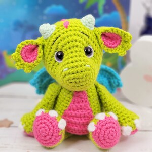 Puede incluir: Un drag&oacute;n de crochet verde con orejas, alas y pies rosas. El drag&oacute;n tiene ojos grandes y negros y una peque&ntilde;a nariz rosa.