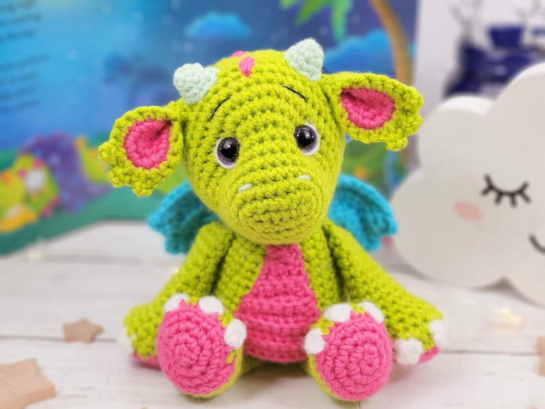 Puede incluir: Un drag&oacute;n de crochet verde con orejas, alas y pies rosas. El drag&oacute;n tiene ojos grandes y negros y una peque&ntilde;a nariz rosa.