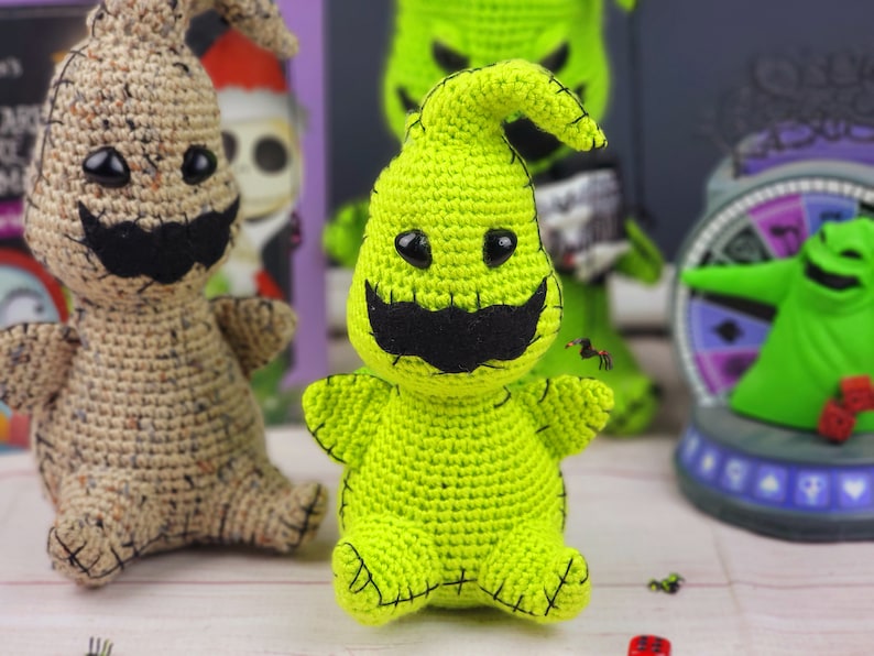 Puede incluir: Dos personajes de fantasmas de crochet verde y beige con ojos negros y un bigote negro. Los fantasmas est&aacute;n sentados sobre una superficie blanca.