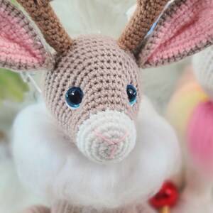 May include: A crocheted reindeer with brown antlers, large blue eyes, and a white fluffy collar. The reindeer's ears are lined with pink. The craft animal is a handmade item.