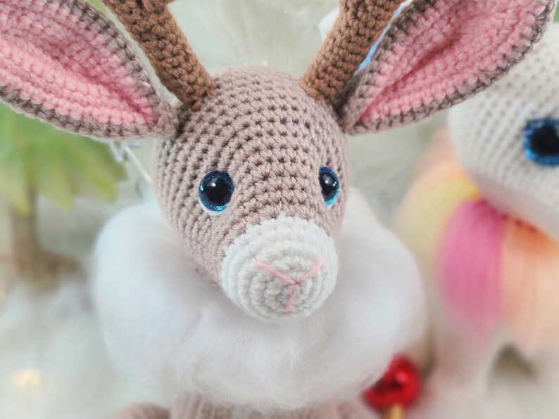 May include: A crocheted reindeer with brown antlers, large blue eyes, and a white fluffy collar. The reindeer's ears are lined with pink. The craft animal is a handmade item.