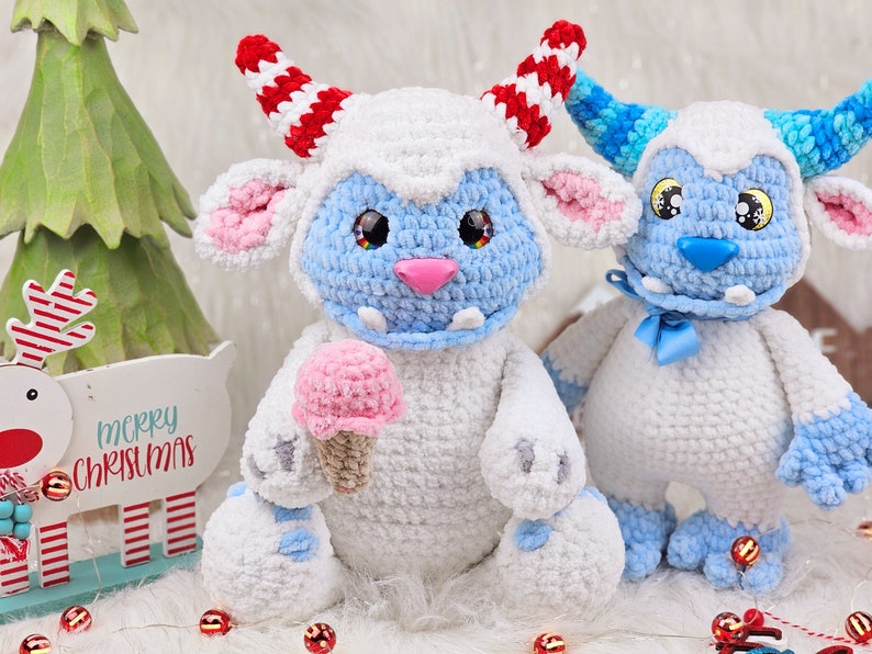 Pu&ograve; includere: Due giocattoli di peluche in crochet bianco con dettagli blu. Un giocattolo ha corna a strisce rosse e bianche e tiene un cono gelato rosa. L'altro giocattolo ha un fiocco blu ed &egrave; seduto accanto al primo giocattolo. I giocattoli sono seduti su una superficie bianca con decorazioni natalizie rosse e bianche.