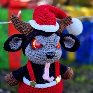 Puede incluir: Un juguete de peluche de crochet con forma de toro que lleva un gorro de Pap&aacute; Noel rojo y blanco. El toro tiene cuernos negros, una cara gris y ojos rojos. El toro lleva un atuendo rojo y negro con un cintur&oacute;n blanco y estrellas doradas.