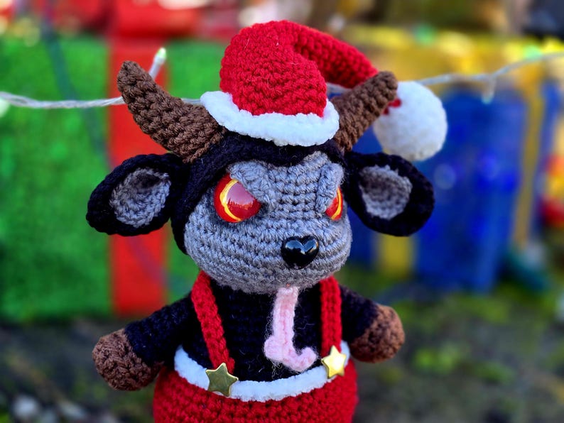 Puede incluir: Un juguete de peluche de crochet con forma de toro que lleva un gorro de Pap&aacute; Noel rojo y blanco. El toro tiene cuernos negros, una cara gris y ojos rojos. El toro lleva un atuendo rojo y negro con un cintur&oacute;n blanco y estrellas doradas.