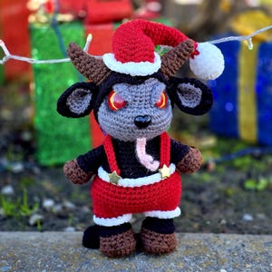 Puede incluir: Un juguete de peluche de crochet con forma de cabra que lleva un gorro de Pap&aacute; Noel rojo y un traje de Pap&aacute; Noel rojo y blanco. El juguete tiene cuernos negros, una cara gris y ojos rojos.