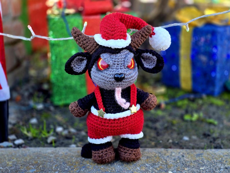 Puede incluir: Un juguete de peluche de crochet con forma de cabra que lleva un gorro de Pap&aacute; Noel rojo y un traje de Pap&aacute; Noel rojo y blanco. El juguete tiene cuernos negros, una cara gris y ojos rojos.