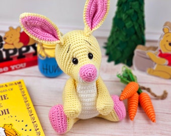 May include: A crocheted yellow bunny rabbit with pink ears and nose. The bunny is sitting on a white wooden surface.