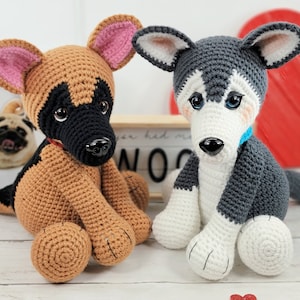 May include: Two crocheted stuffed dog toys. One is a brown and black German Shepherd with pink ears. The other is a gray and white Husky with blue eyes. Both dogs are sitting on a white wooden surface.