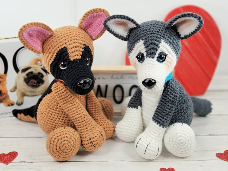 May include: Two crocheted stuffed dog toys. One is a brown and black German Shepherd with pink ears. The other is a gray and white Husky with blue eyes. Both dogs are sitting on a white wooden surface.