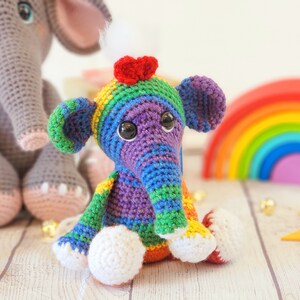 May include: A colorful crocheted elephant toy with a rainbow striped body and a red heart on its head. The elephant is sitting on a white wooden surface.