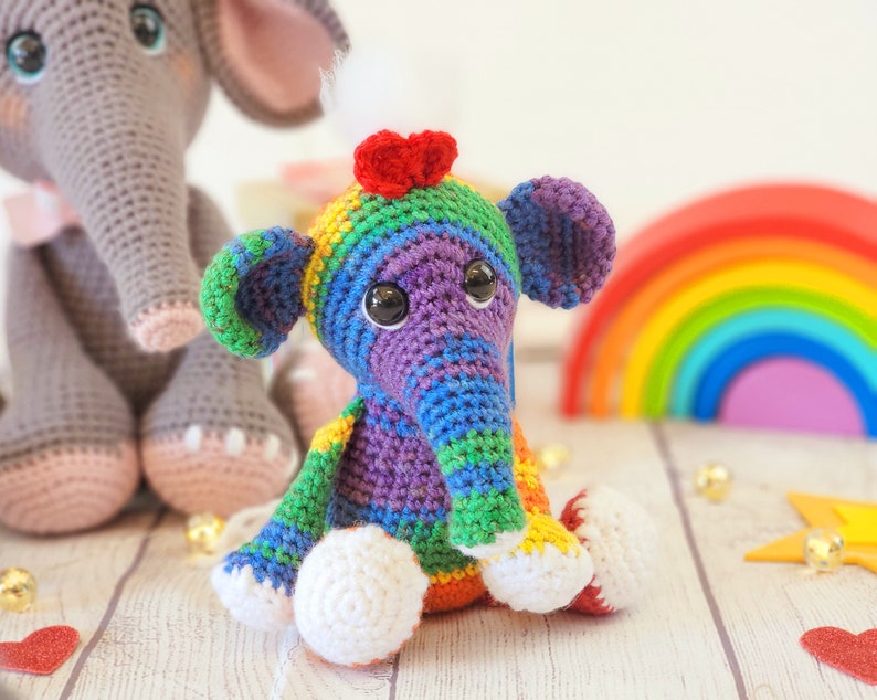 May include: A colorful crocheted elephant toy with a rainbow striped body and a red heart on its head. The elephant is sitting on a white wooden surface.