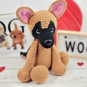 May include: A crocheted German Shepherd puppy toy with brown and black fur, pink ears, and black eyes. The toy is sitting on a white wooden surface with a sign that says "Woof" in black letters.