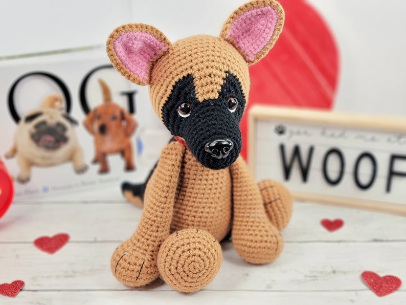 May include: A crocheted German Shepherd puppy toy with brown and black fur, pink ears, and black eyes. The toy is sitting on a white wooden surface with a sign that says "Woof" in black letters.