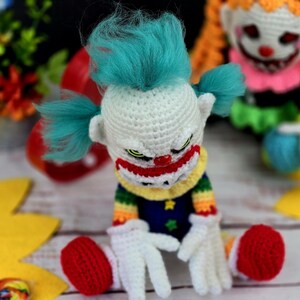May include: A crocheted clown doll with a white face, red nose, and blue hair. The doll is wearing a rainbow striped shirt and blue trousers with stars. The doll is sitting on a white surface with yellow and brown decorations.