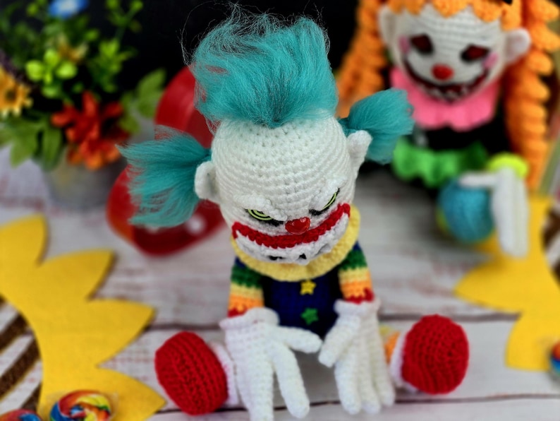 May include: A crocheted clown doll with a white face, red nose, and blue hair. The doll is wearing a rainbow striped shirt and blue trousers with stars. The doll is sitting on a white surface with yellow and brown decorations.