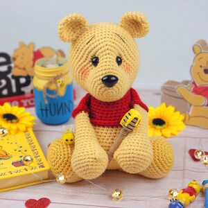 K&ouml;nnte beinhalten: Ein geh&auml;kelter Winnie Puuh B&auml;r, der ein rotes Hemd tr&auml;gt und einen Honigl&ouml;ffel h&auml;lt. Der B&auml;r sitzt auf einer Holzoberfl&auml;che mit einem Buch, einem Honigglas und einer Sonnenblume.