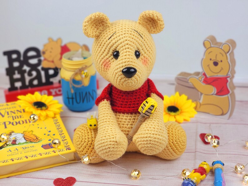 K&ouml;nnte beinhalten: Ein geh&auml;kelter Winnie Puuh B&auml;r, der ein rotes Hemd tr&auml;gt und einen Honigl&ouml;ffel h&auml;lt. Der B&auml;r sitzt auf einer Holzoberfl&auml;che mit einem Buch, einem Honigglas und einer Sonnenblume.