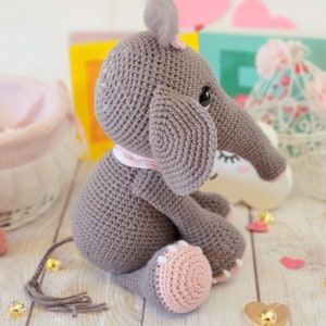 May include: A gray crocheted elephant toy with pink accents, sitting on a wooden surface. The elephant has a long trunk and large ears.
