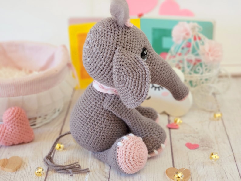 May include: A gray crocheted elephant toy with pink accents, sitting on a wooden surface. The elephant has a long trunk and large ears.