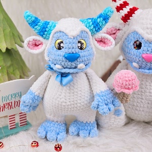 Pu&ograve; includere: Due giocattoli di peluche all'uncinetto a forma di mostri yeti blu e bianchi. Un yeti tiene un cono gelato rosa all'uncinetto. I mostri yeti sono seduti su una superficie bianca e soffice.