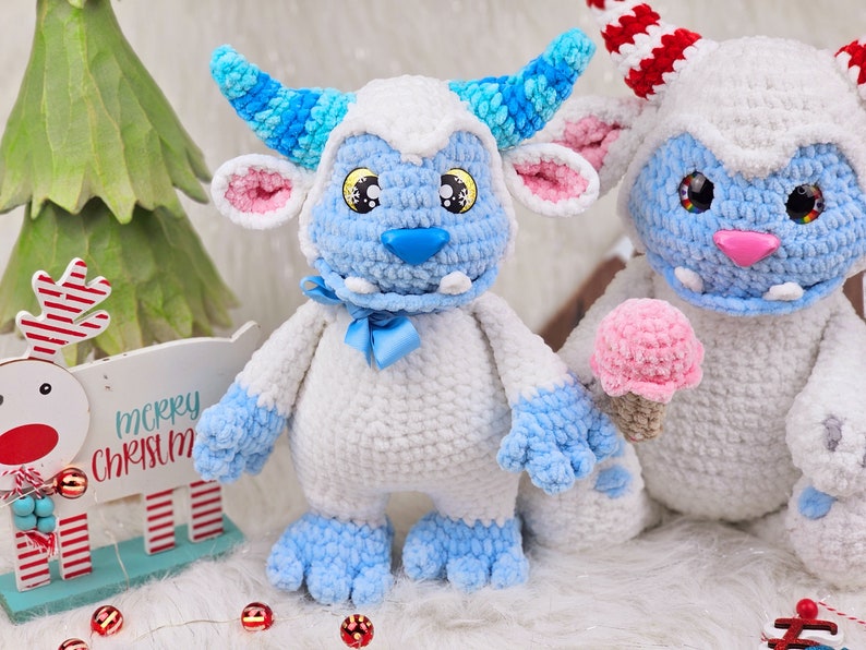 Pu&ograve; includere: Due giocattoli di peluche all'uncinetto a forma di mostri yeti blu e bianchi. Un yeti tiene un cono gelato rosa all'uncinetto. I mostri yeti sono seduti su una superficie bianca e soffice.
