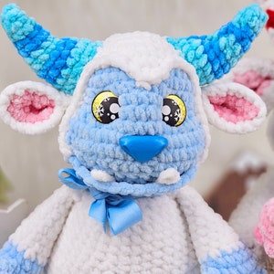 Pu&ograve; includere: Un giocattolo di peluche blu e bianco lavorato a uncinetto con naso e orecchie rosa. Il giocattolo ha un fiocco blu intorno al collo e occhi gialli con fiocchi di neve.