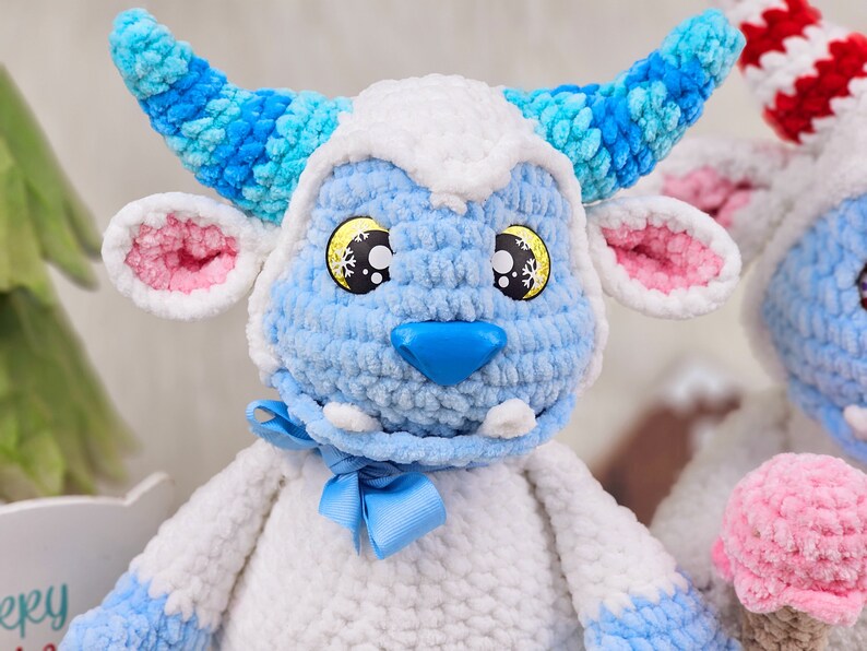 Pu&ograve; includere: Un giocattolo di peluche blu e bianco lavorato a uncinetto con naso e orecchie rosa. Il giocattolo ha un fiocco blu intorno al collo e occhi gialli con fiocchi di neve.