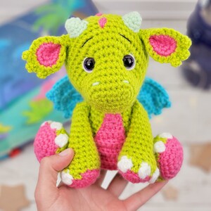 May include: A green crocheted dragon with pink ears, blue wings, and white spots on its feet. The dragon is sitting on a hand.