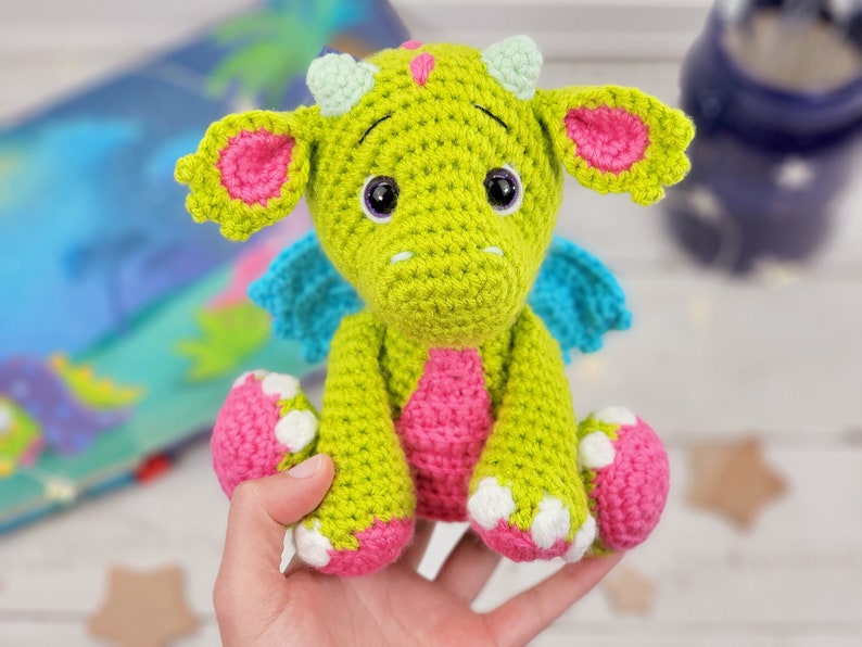 May include: A green crocheted dragon with pink ears, blue wings, and white spots on its feet. The dragon is sitting on a hand.