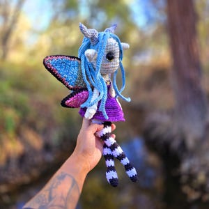 May include: A crocheted fairy doll with blue hair, purple dress, and black and white striped legs. The fairy has wings with blue, pink, and silver accents. The doll is being held by a hand with a tattoo.