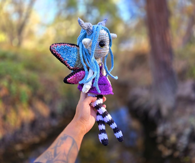 May include: A crocheted fairy doll with blue hair, purple dress, and black and white striped legs. The fairy has wings with blue, pink, and silver accents. The doll is being held by a hand with a tattoo.