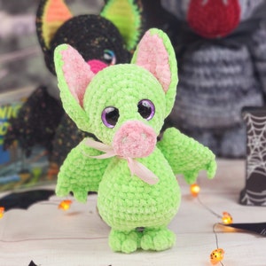 May include: A green crocheted bat with pink ears and a pink nose. The bat has large purple eyes and a pink ribbon around its neck. The bat is sitting on a white surface with a black and white bat wing in the background.