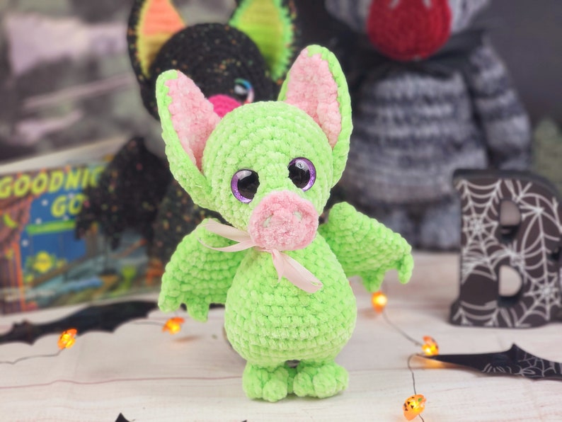 May include: A green crocheted bat with pink ears and a pink nose. The bat has large purple eyes and a pink ribbon around its neck. The bat is sitting on a white surface with a black and white bat wing in the background.