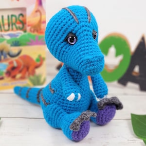Puede incluir: Un juguete de dinosaurio de crochet azul con detalles en morado y gris. El dinosaurio está sentado sobre una superficie blanca con hojas verdes en el fondo.