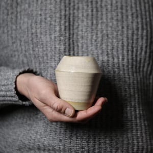 Könnte beinhalten: Eine handgefertigte, cremefarbene Keramikvase mit einer einzigartigen, geometrischen Form. Die Vase hat eine hellbeige Farbe mit leicht strukturierter Oberfläche und einem hellgelben Boden. Die Vase wird in der Hand einer Person vor einem grauen Pullover gehalten.