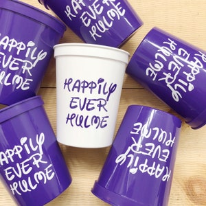 May include: Six purple and white plastic cups with the text "Happily Ever Hulme" written on them. The cups are arranged on a wooden surface.