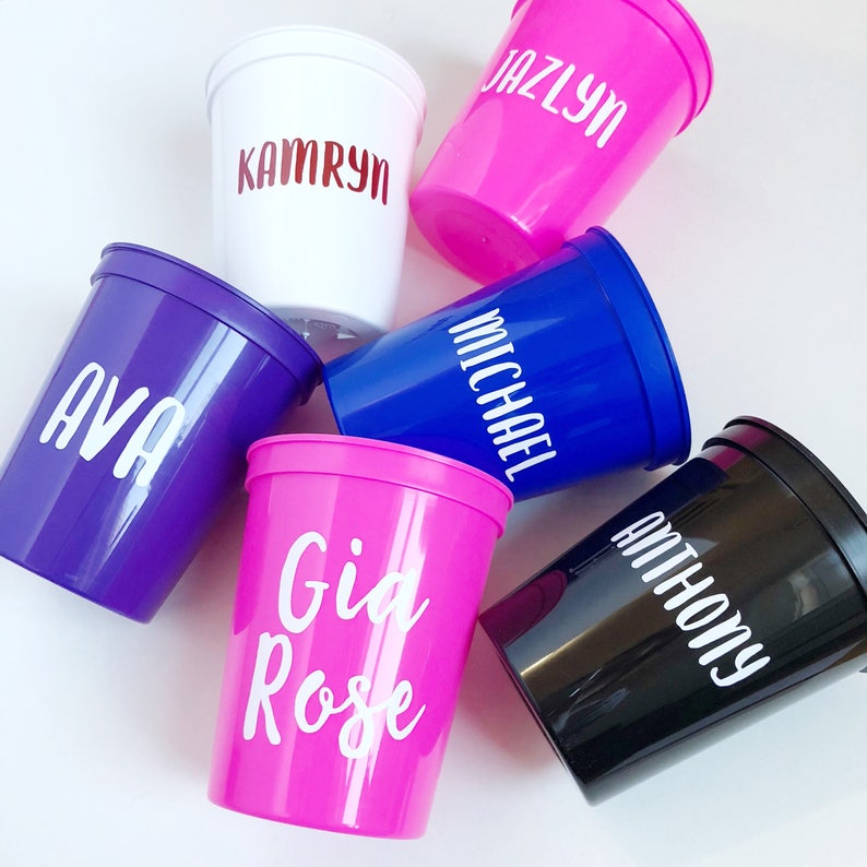 May include: Six personalized plastic cups with different names printed on them. The cups are in various colors, including white, pink, purple, blue, and black.