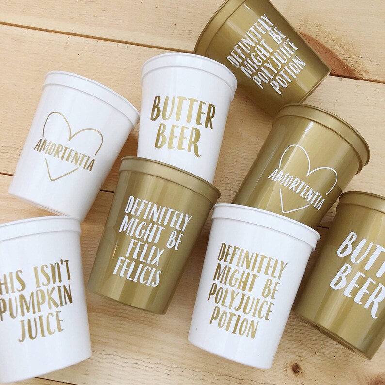 May include: Eight gold and white plastic cups with various Harry Potter themed phrases printed on them. The phrases include "Butter Beer", "Amortentia", "Definitely Might Be Polyjuice Potion", "This Isn't Pumpkin Juice", and "Felix Felicis".