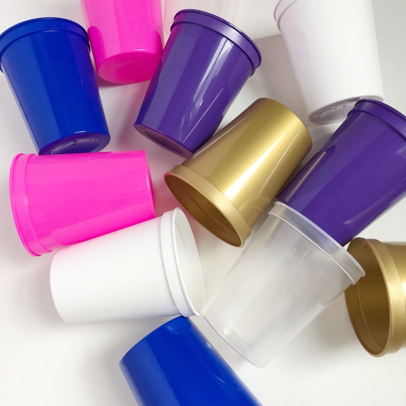 May include: A collection of colorful plastic cups in various shades of blue, purple, pink, gold, and white. The cups are arranged in a random pattern on a white surface.