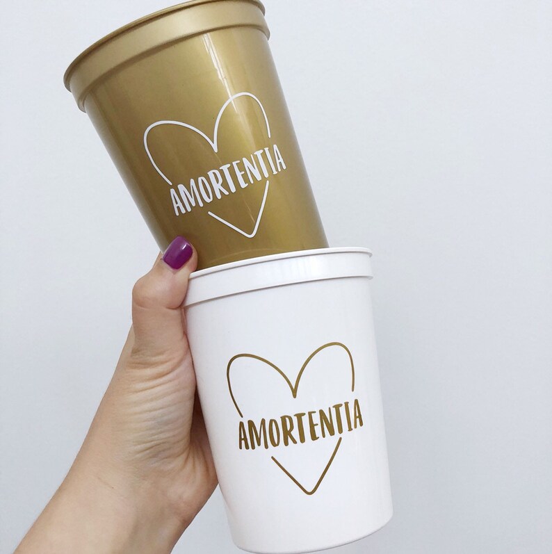 May include: Two plastic cups with a white heart design and the word "Amortentia" written inside the heart. The top cup is gold and the bottom cup is white.