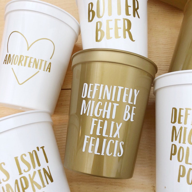 May include: A collection of six white and gold plastic cups with various phrases written on them in gold lettering. The phrases include "Amortentia", "Butter Beer", "Definitely Might Be Felix Felicis", and "S Isn't Pumpkin".
