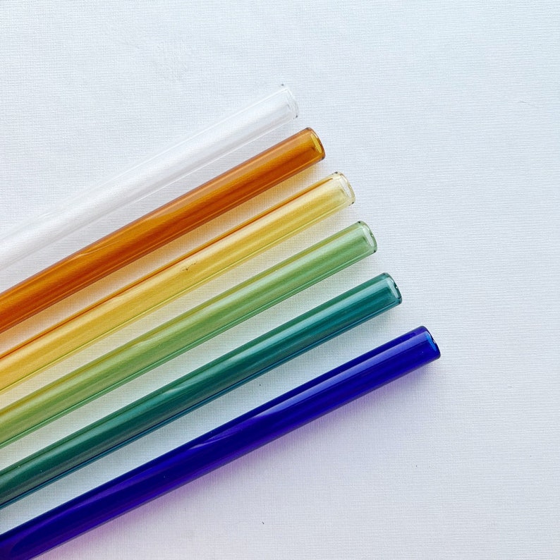 May include: Six glass drinking straws in various colours, including clear, orange, yellow, green, teal, and blue. The straws are arranged in a rainbow pattern, with the clear straw at the top and the blue straw at the bottom.