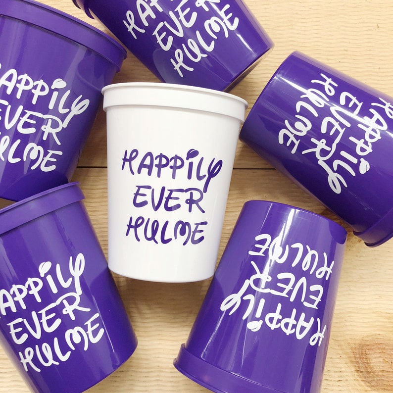 May include: Six purple and white plastic cups with the text "Happily Ever Hulme" written on them. The cups are arranged on a wooden surface.