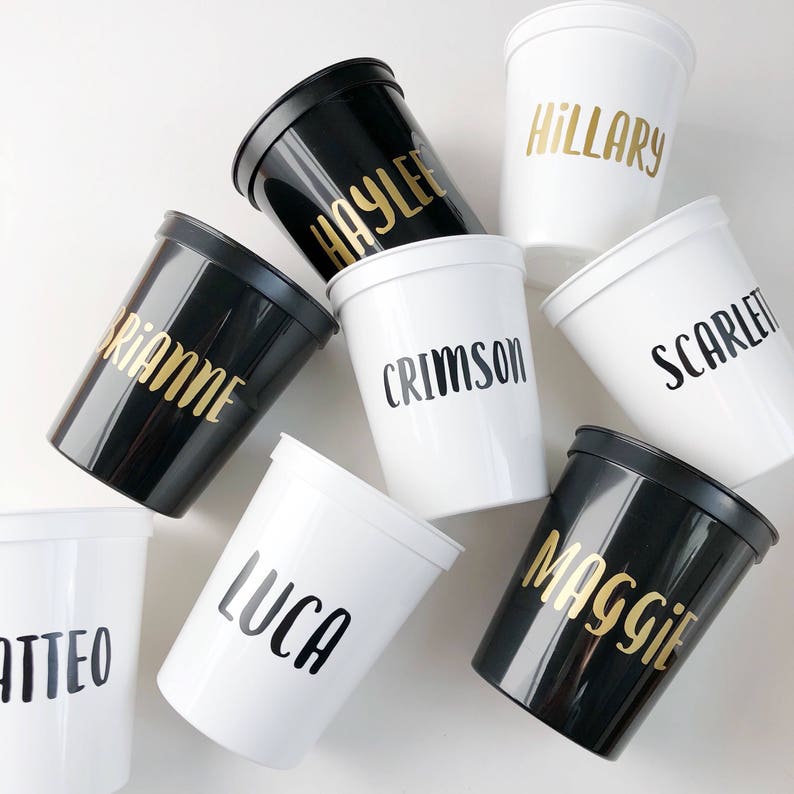 May include: Nine black and white plastic cups with gold lettering. The cups are labeled with the names: Brianne, Haylee, Crimson, Hillary, Scarlett, Matteo, Lucca, Maggie.