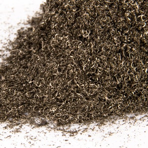 May include: A pile of metal shavings, a byproduct of metalworking, on a white background. The shavings are a dark gray color and are tightly packed together.