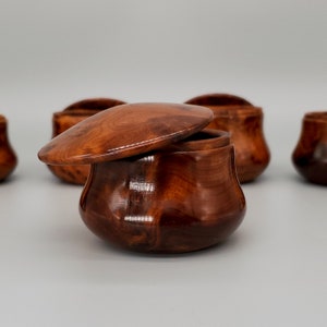 May include: A set of five wooden bowls with lids. The bowls are a rich brown color and have a smooth, polished finish. The lids are slightly domed and fit snugly on top of the bowls.