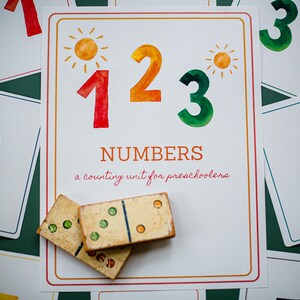 Puede incluir: Una tarjeta blanca con los números 1, 2 y 3 en rojo, naranja y verde, con ilustraciones de sol. El texto "NUMBERS a counting unit for preschoolers" está debajo. Dos dominós en primer plano.